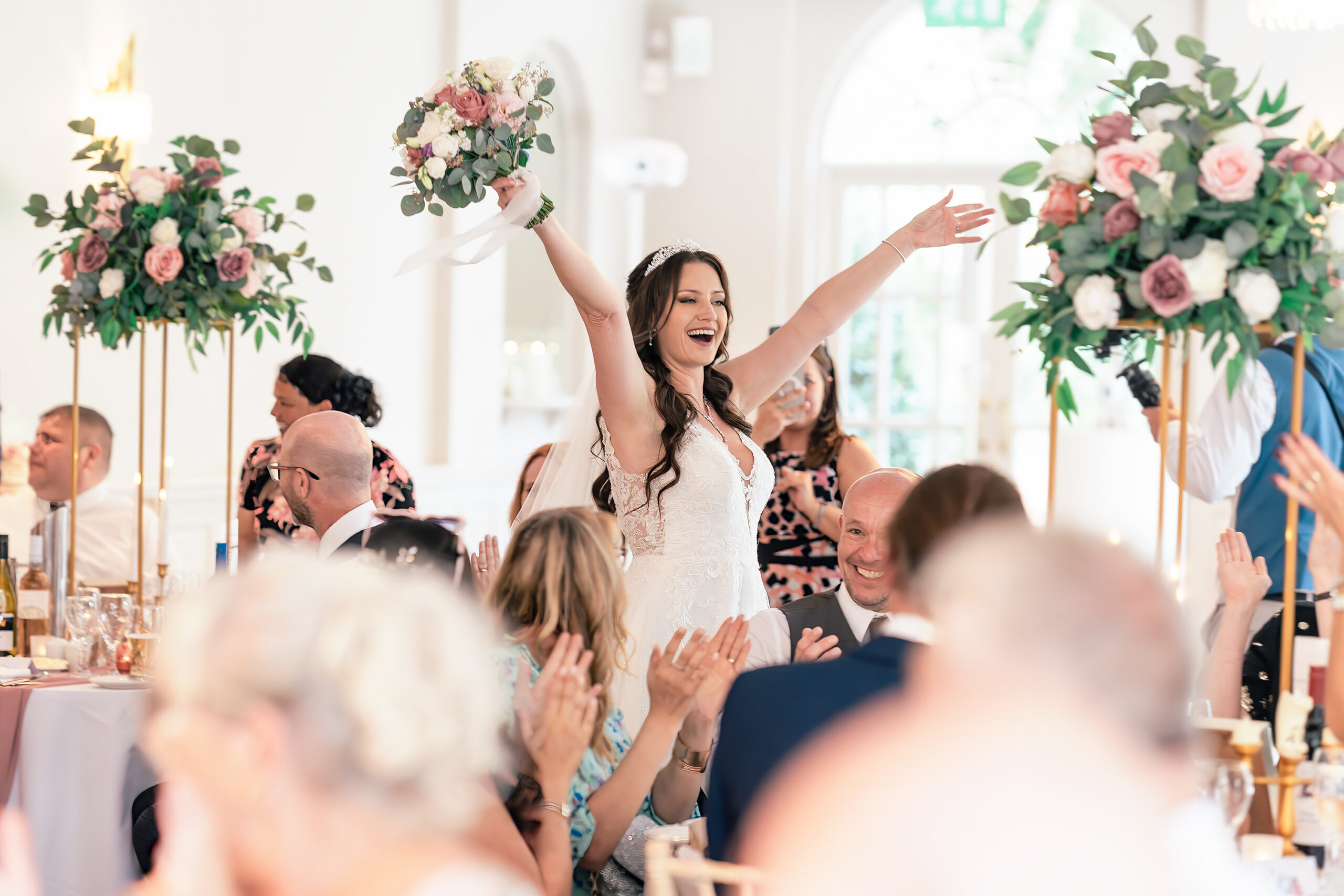 A bride in a white gown raises her arms joyfully while holding a bouquet, walking among seated guests at a wedding reception. The room is decorated with tall floral arrangements, and the guests are clapping and smiling, creating a festive atmosphere. The Orangery, Maidstone, Kent.