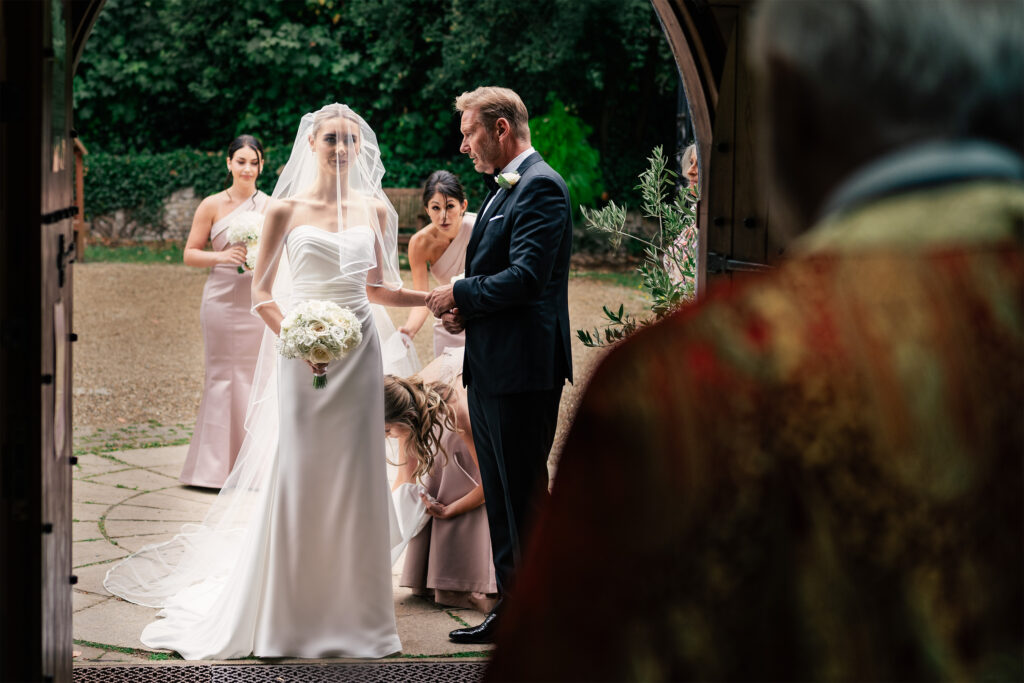 A bride and groom stand together at the entrance of a church during their wedding ceremony. The bride, wearing a veil and holding a bouquet, is accompanied by bridesmaids adjusting her dress. The groom, in a black suit, holds her hand. A priest stands in the foreground. Sopwell House Wedding.