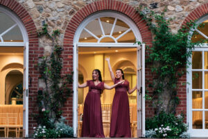 Bridesmaids, in matching burgundy dresses stand joyfully in a doorway framed by brick and greenery. They pose with arms raised excitedly. The background features an elegant interior with arched windows and light wooden chairs. The Orangery, Maidstone, Kent.