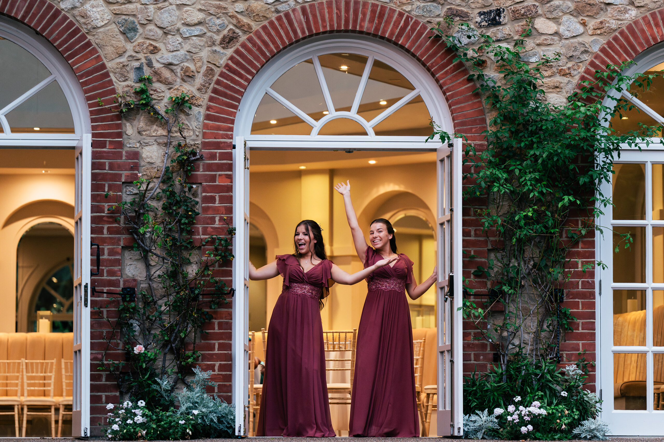 Bridesmaids, in matching burgundy dresses stand joyfully in a doorway framed by brick and greenery. They pose with arms raised excitedly. The background features an elegant interior with arched windows and light wooden chairs. The Orangery, Maidstone, Kent.