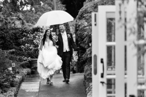 A joyful bride and groom walk down a garden path under an umbrella. The bride holds up her dress, and the groom, in a suit, holds the umbrella over them. The scene is surrounded by lush greenery and an open door is visible in the foreground. The image is in black and white. The Orangery, Maidstone, Kent.