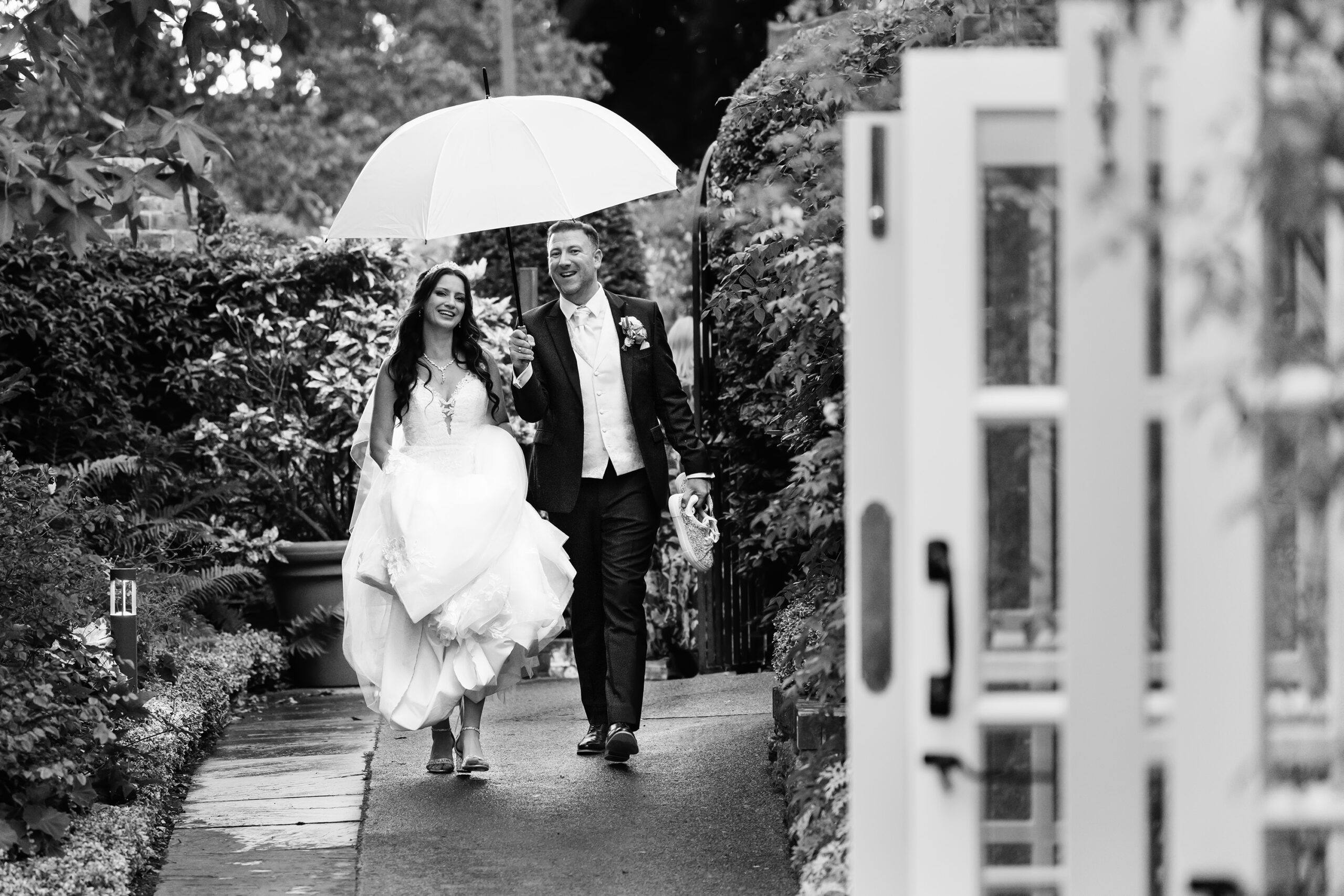 A joyful bride and groom walk down a garden path under an umbrella. The bride holds up her dress, and the groom, in a suit, holds the umbrella over them. The scene is surrounded by lush greenery and an open door is visible in the foreground. The image is in black and white. The Orangery, Maidstone, Kent.