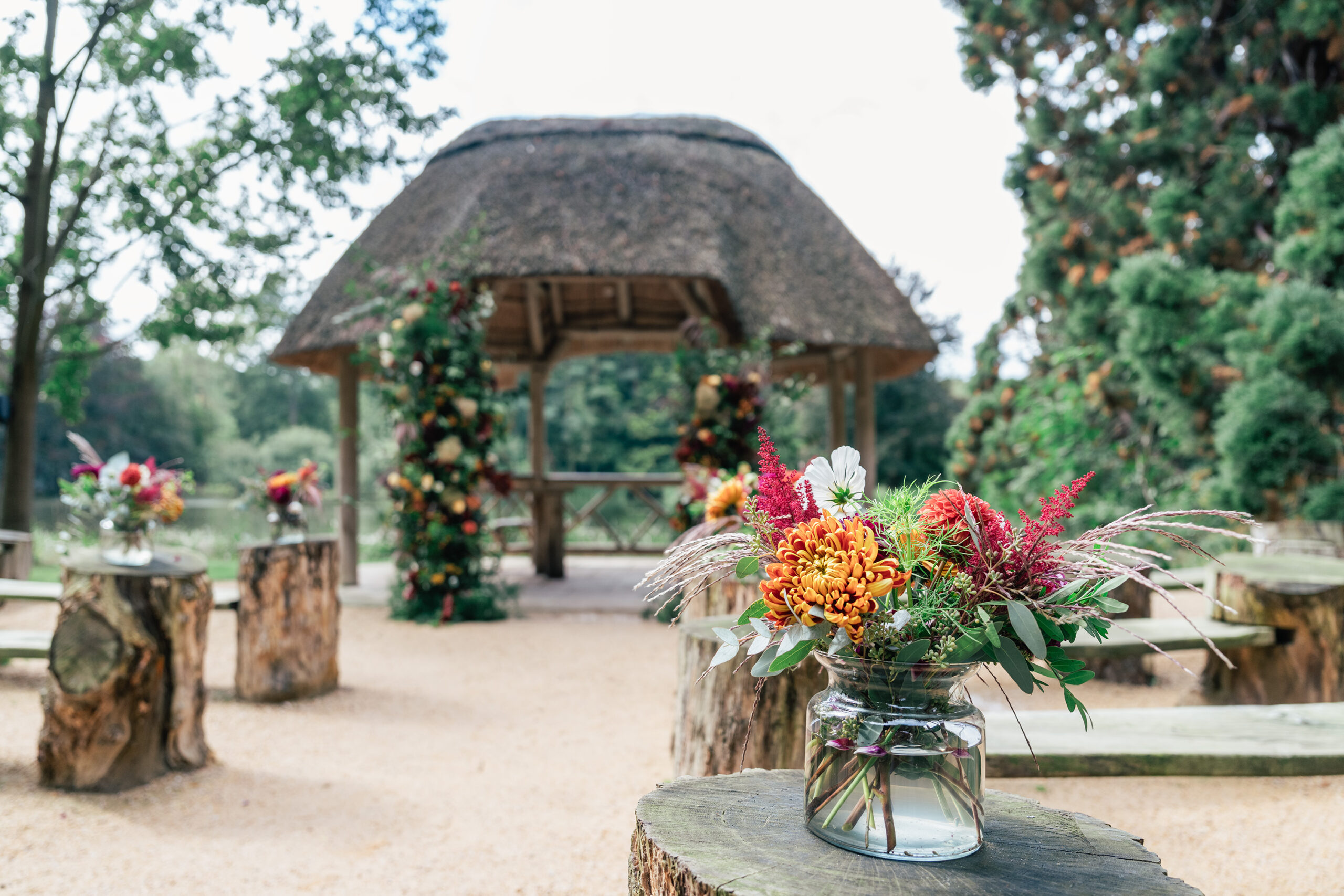 An outdoor wedding setup with a small thatched-roof gazebo in the background, decorated with vibrant floral arrangements. Wooden logs serve as benches, and jarred floral centerpieces are placed on the logs. Lush greenery surrounds the area. The Orangery, Maidstone, Kent.