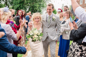 A bride and groom, both smiling and dressed in wedding attire, walk arm in arm down an outdoor pathway as guests cheer and throw confetti. The bride holds a bouquet of white flowers. The scene is joyful and celebratory, with greenery in the background.