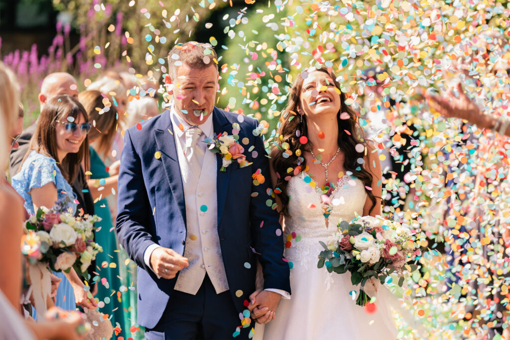 A joyful bride and groom walk hand in hand, smiling as colorful confetti rains down around them. The groom wears a blue suit and beige vest, while the bride is in a white dress holding a bouquet. Guests with flowers applaud in the background.