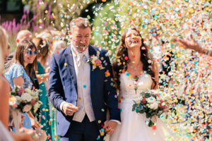 A joyful bride and groom walk hand in hand, smiling as colorful confetti rains down around them. The groom wears a blue suit and beige vest, while the bride is in a white dress holding a bouquet. Guests with flowers applaud in the background.