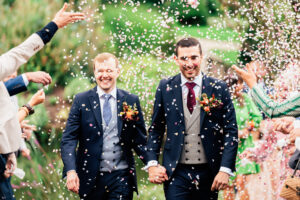 Two grooms in suits, one with a navy suit and red tie, the other with a navy suit and light grey tie, walk hand in hand while smiling under a shower of confetti thrown by guests, celebrating their wedding. They are surrounded by greenery in an outdoor setting. The Orangery, Kent