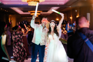 A couple dressed in formal attire, both wearing glowing glasses, joyfully wave glowing sticks in the air at a wedding reception. Other guests, some with glowing accessories, dance and celebrate in a dimly lit, elegant venue with chandeliers and festive lighting. The Orangery, Maidstone, Kent.