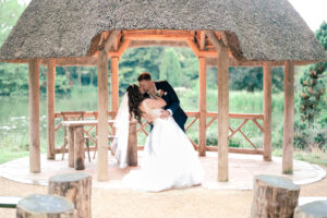 A newlywed couple shares a romantic kiss under a thatched gazebo by a serene lake. The bride, in a white gown, holds her bouquet while leaning back, supported by the groom in a navy blue suit. Surrounding them are lush greenery and wooden elements.