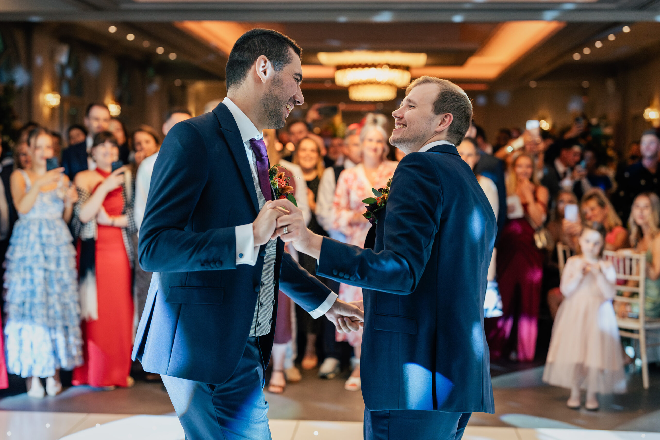 Two grooms in formal wear dance joyfully at their wedding reception. Onlookers, including family and friends, smile and clap as they watch. The background features a warmly lit, elegant venue filled with guests in festive attire, creating a celebratory atmosphere. The Orangery, Maidstone, Kent.