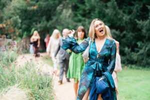 A woman in a green, leaf-patterned dress enthusiastically points to something off-camera, her mouth wide open in excitement. She stands on a garden path with several other people blurred in the background, surrounded by greenery and tall plants. The Orangery, Kent