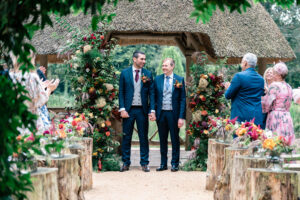 A joyful wedding scene with two grooms holding hands and smiling under a rustic, thatched-roof archway adorned with colorful flowers and greenery. Guests cheer and clap around them in celebration. The ceremony takes place outdoors, surrounded by nature. The Orangery, Maidstone, Kent.