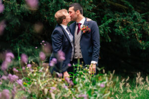 Two men, dressed in suits, share a kiss in an outdoor setting surrounded by lush greenery and purple flowers. One man wears a boutonniere and the other sports a vest and tie. The scene conveys warmth and affection amidst a natural backdrop.The Orangery, Kent