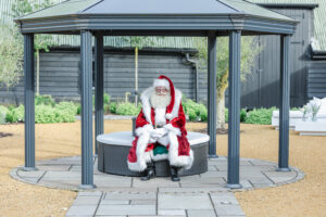 A person dressed as Santa Claus sits on a white circular bench under a gazebo. The background shows a garden area with plants and a pathway, and a dark wooden building. Santa is wearing a red suit with white fur trim, black boots, and a red hat.