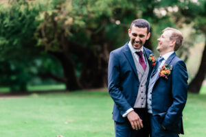 Two men in suits stand close together outdoors, smiling and laughing. Both wear dark suits with colorful boutonnieres, and one has a maroon tie, while the other is in a blue tie. The background features lush green, blurred trees and grass. The Orangery, Kent