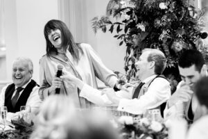 A black and white photo of a lively wedding scene. A woman with long hair joyfully holds a microphone, laughing, as she stands next to a seated, smiling man dressed in formal attire. They are surrounded by guests who are also laughing, with floral decorations in the background. The Orangery, Kent