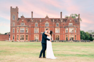 A couple in formal attire embrace lovingly in front of a large, historic brick mansion at sunset. The bride is wearing a white wedding dress, and the groom is in a dark suit. The sky is painted with soft pink and blue hues, adding to the romantic atmosphere. Hertfordshire Golf & Country Club.