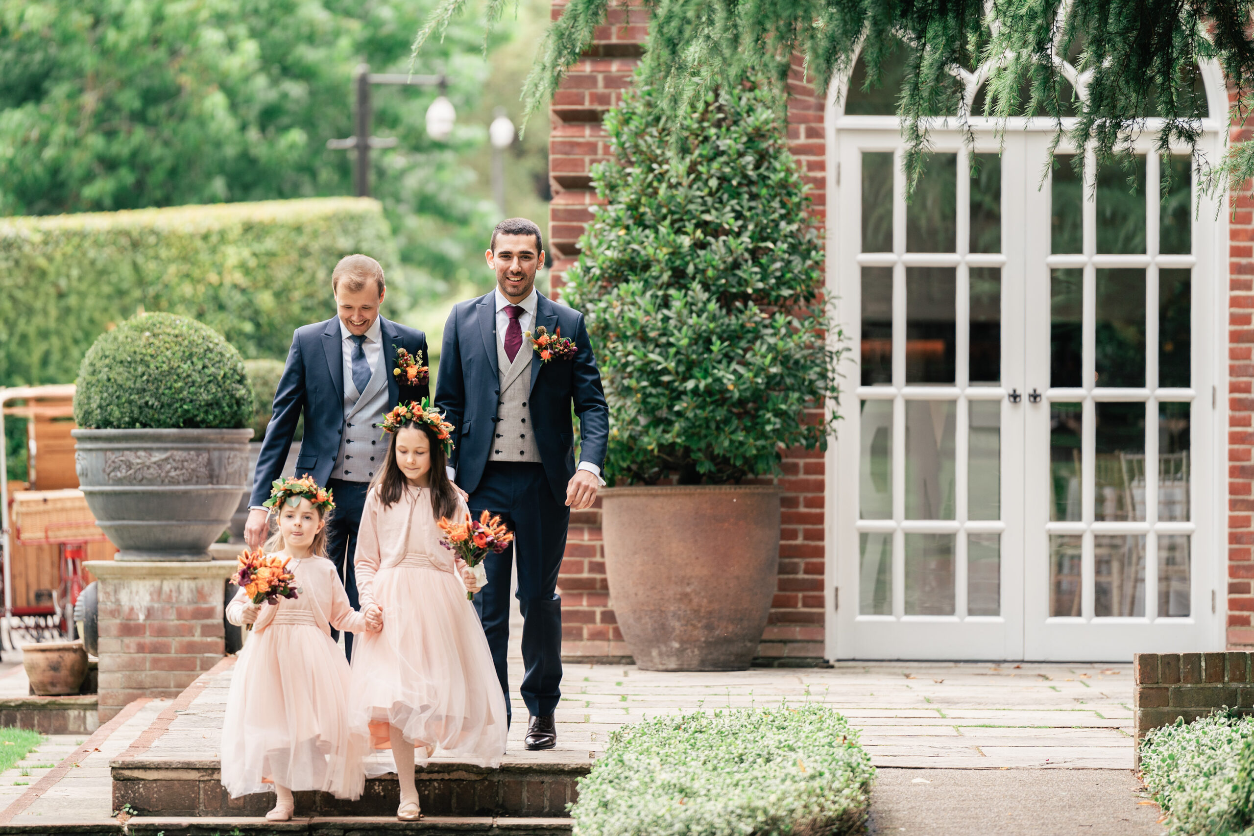 Two men in suits, both smiling, walk down steps with two girls in light pink dresses and floral crowns. The men hold bouquets, one hand on each girl's shoulder. Behind them is a brick building with a white door, large potted plants, and green hedges. The Orangery, Kent