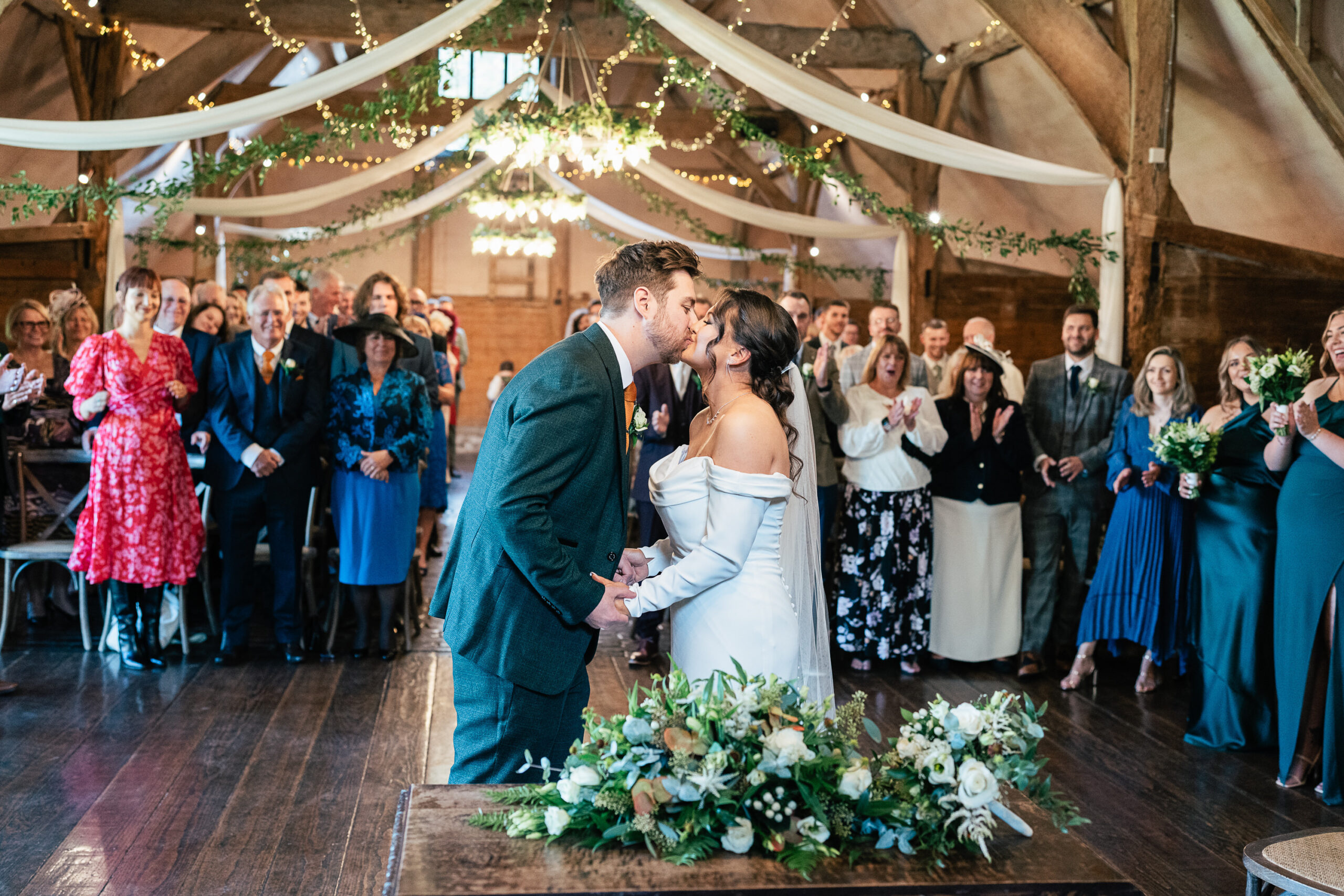 A bride and groom share a kiss at their wedding ceremony in a rustic venue decorated with draped fabric and greenery. Guests stand and applaud, with some taking photos. Floral arrangements adorn the scene.