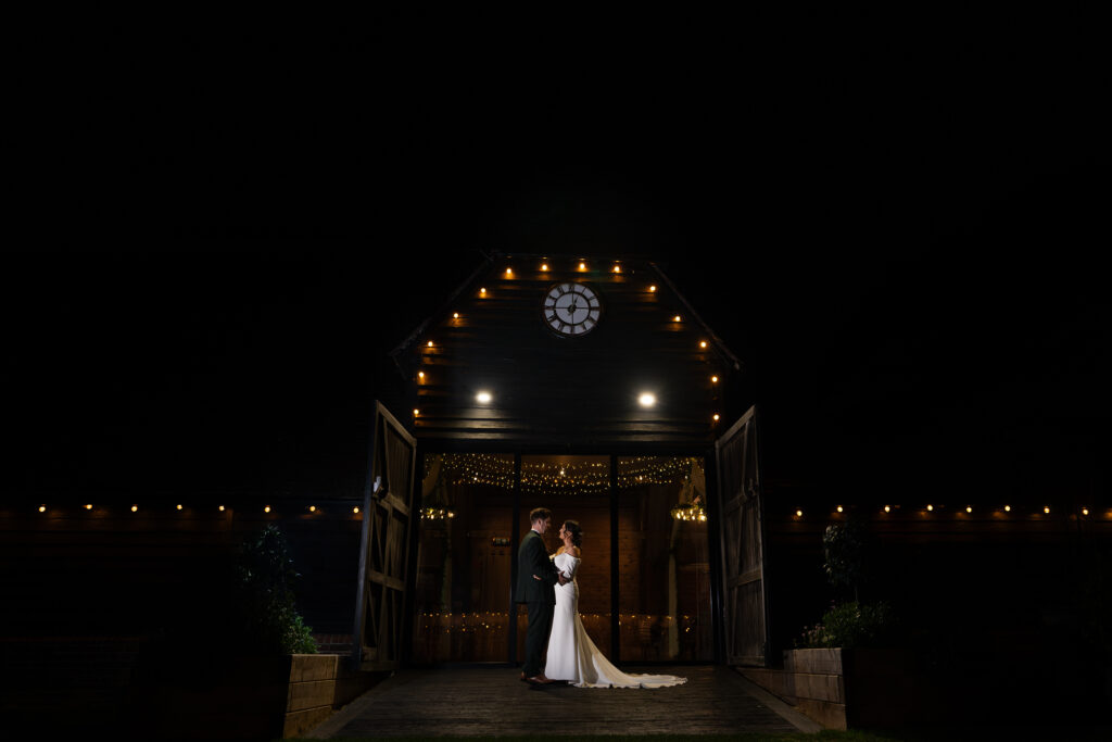 A bride and groom stand facing each other at night in front of a rustic barn, illuminated by strings of lights. The barn doors are open, revealing a warmly lit interior adorned with more lights. The sky is dark, highlighting the cozy ambiance. Lains Barn, Oxfordshire