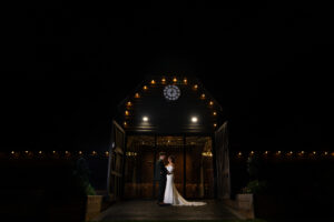 A bride and groom stand facing each other at night in front of a rustic barn, illuminated by strings of lights. The barn doors are open, revealing a warmly lit interior adorned with more lights. The sky is dark, highlighting the cozy ambiance. Lains Barn, Oxfordshire