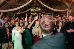 A joyous group of wedding guests, dressed in formal attire, sing and cheer with enthusiasm beneath strings of lights and wooden beams. They are inside a rustic venue, creating a festive and lively atmosphere. Lains Barn, Oxfordshire