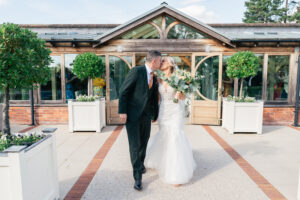 A newlywed couple kisses in front of a rustic building with large glass windows. The bride wears a white gown and holds a bouquet, while the groom is in a black suit. They are walking on a path lined with large potted plants. Gaynes Park, Essex.