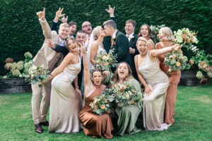 A joyful wedding party posed on grass, smiling and celebrating. The group is dressed in elegant suits and dresses, with the bridesmaids holding bouquets. A hedge serves as the backdrop. Gaynes Park, Essex.