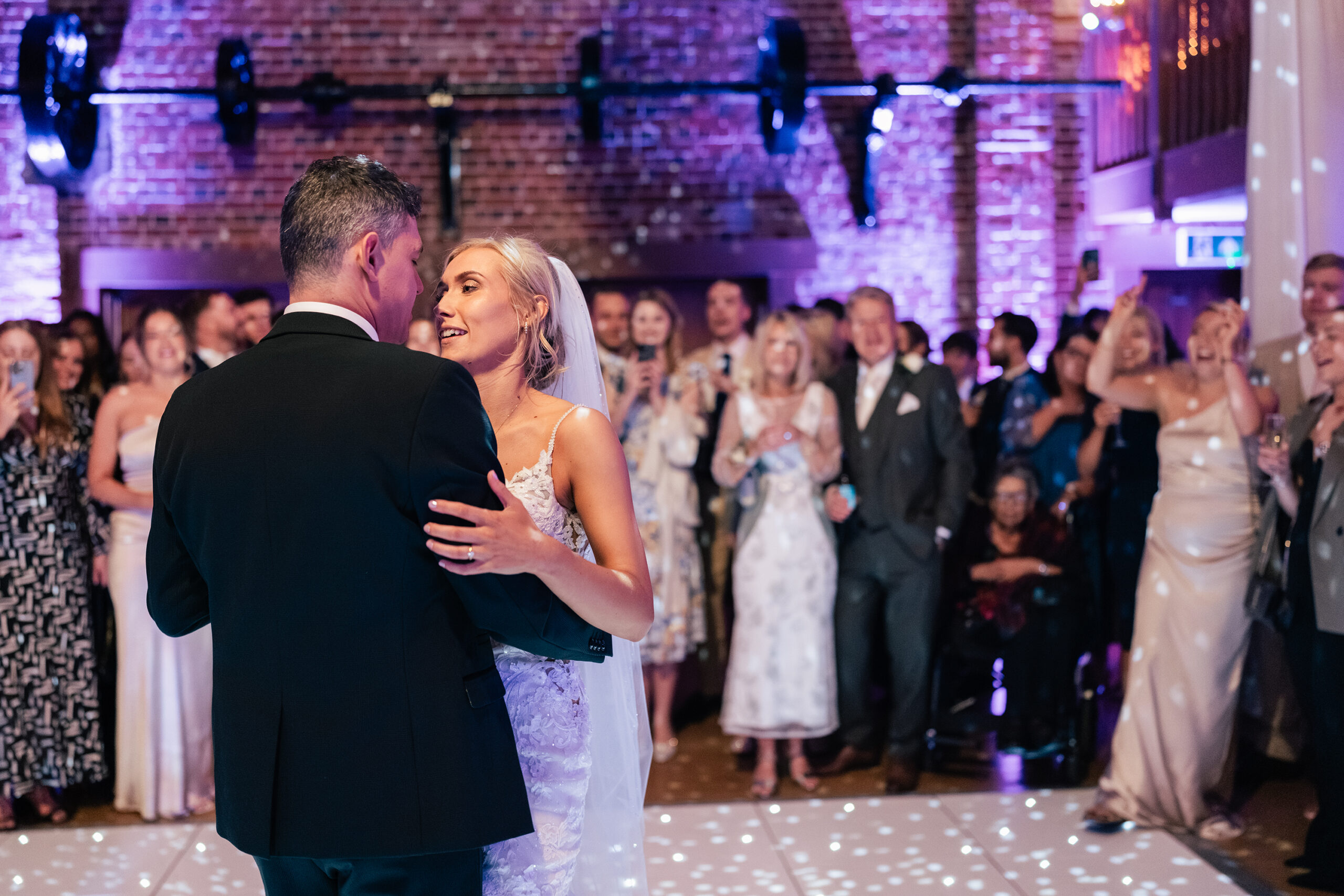 A bride and groom share their first dance at a wedding reception. They are surrounded by applauding guests. The venue features exposed brick walls and purple lighting, with a DJ setup and dotted light patterns on the floor. Gaynes Park, Essex.