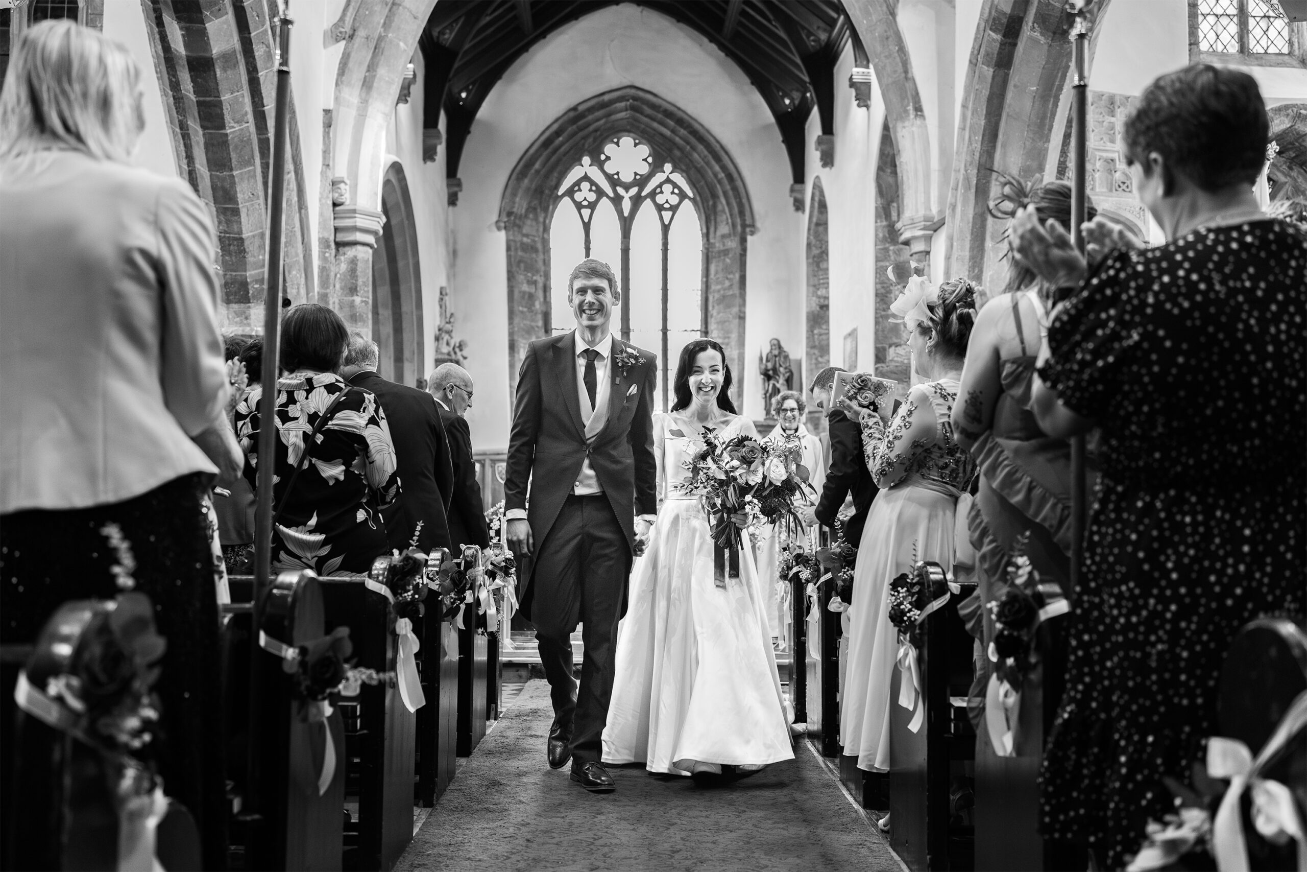 A bride and groom walk down the aisle of a church, smiling and holding hands. Guests stand and applaud on either side. The couple is dressed in formal attire, and the church features stained glass windows and wooden pews. Black and white photo.