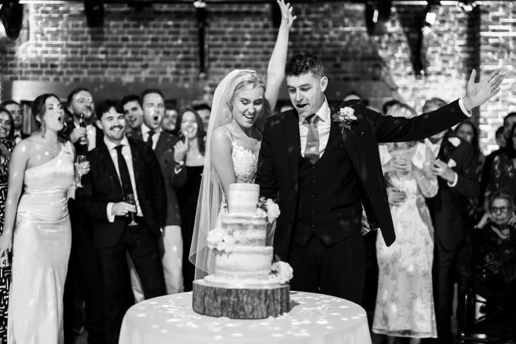 A bride and groom enthusiastically cut a tiered wedding cake while surrounded by a cheering crowd. The black and white photo captures the joyous celebration in a festive, brick-walled venue. Gaynes Park, Essex.