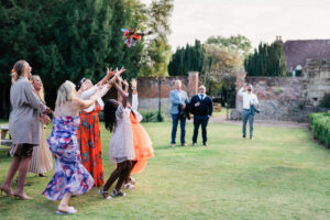 A group of excited women and girls reach to catch a bouquet in a garden setting during a wedding. Onlookers in the background watch and take photos. Brick walls and greenery surround the scene, creating a lively, festive atmosphere.