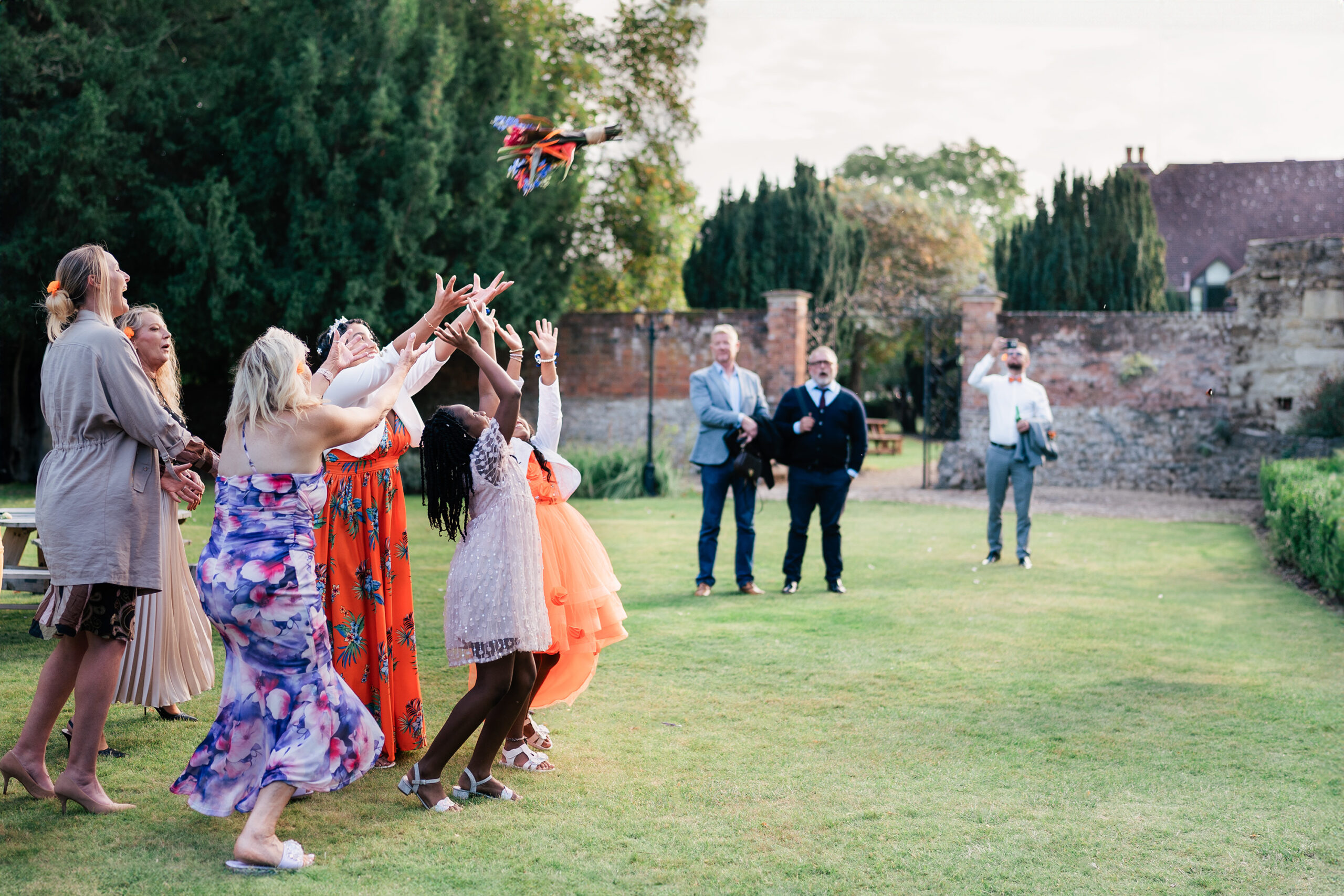 A group of excited women and girls reach to catch a bouquet in a garden setting during a wedding. Onlookers in the background watch and take photos. Brick walls and greenery surround the scene, creating a lively, festive atmosphere.