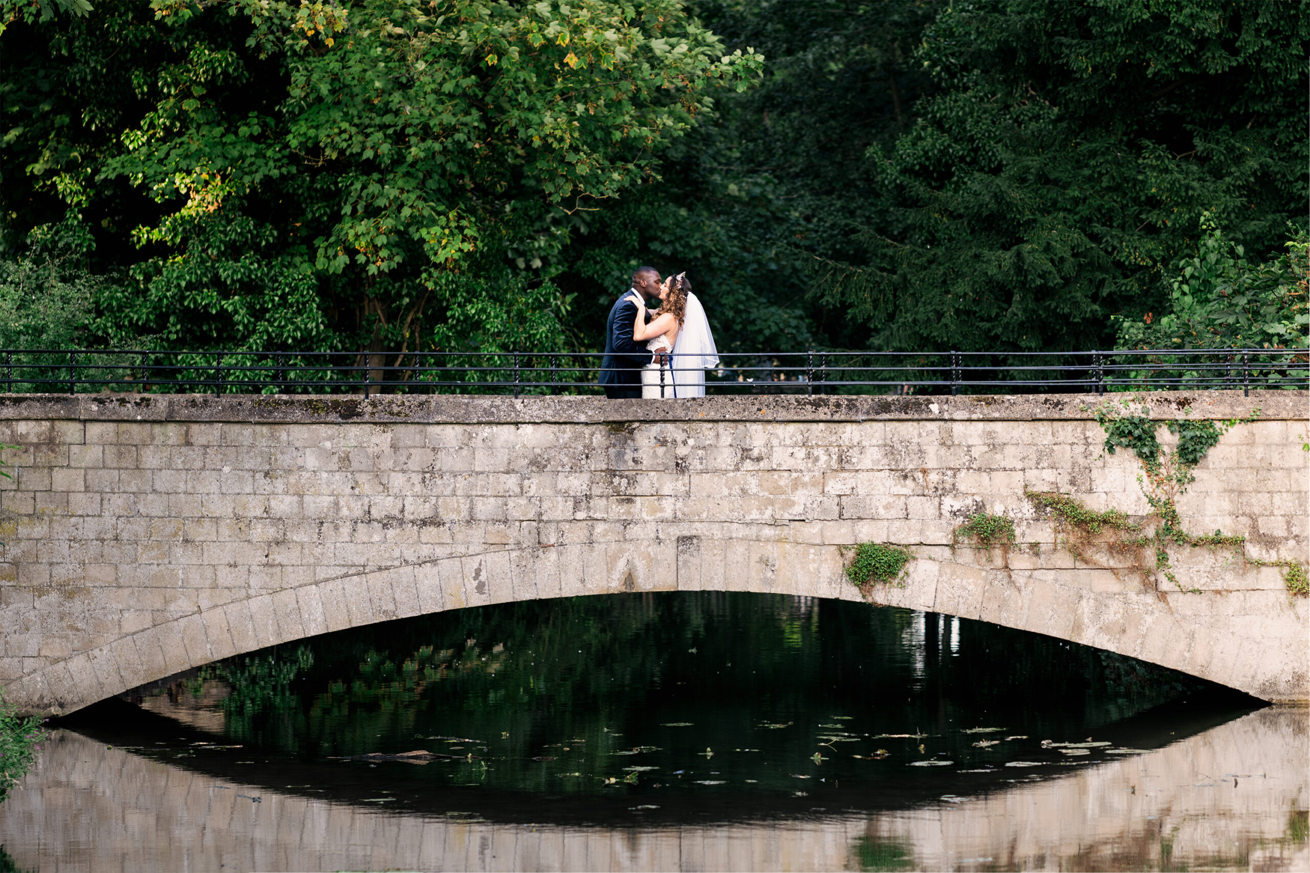 A bride and groom share a kiss on a stone bridge surrounded by lush greenery, reflected in the calm water below. The scene is serene and romantic. Coseners House, Oxfordshire