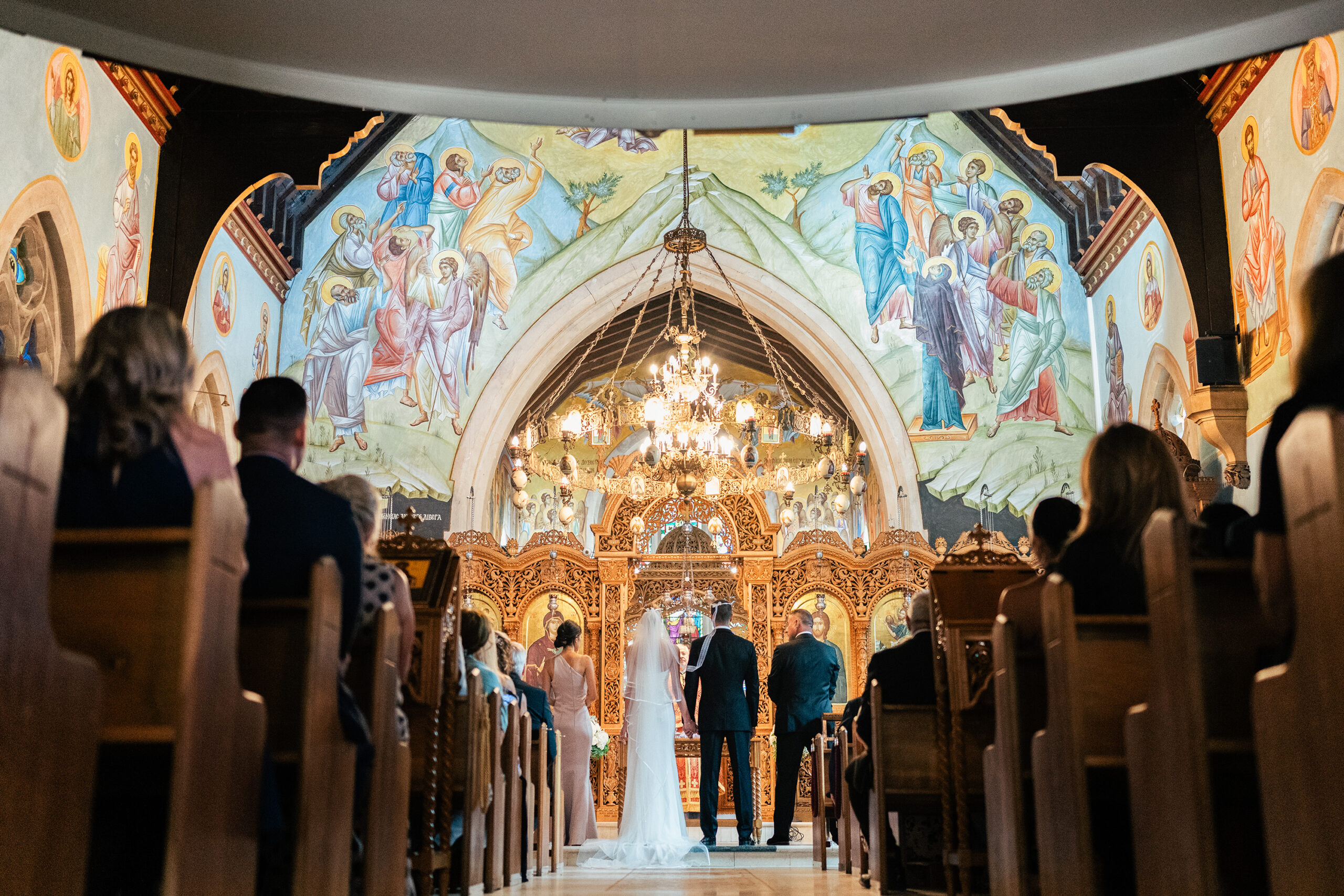 A couple stands at the altar in a richly decorated church with colorful frescoes and ornate chandeliers. Guests sit in wooden pews, observing the ceremony. The vaulted ceiling features vibrant religious artwork.