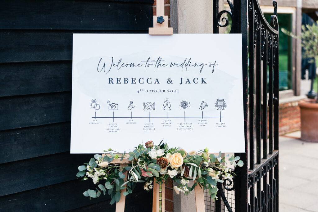 A wedding sign on an easel reads "Welcome to the wedding of Rebecca & Jack, 4th October 2024." It includes icons representing events like drinks, dinner, and dancing. The sign is decorated with a floral arrangement at the bottom. Gaynes Park, Essex.