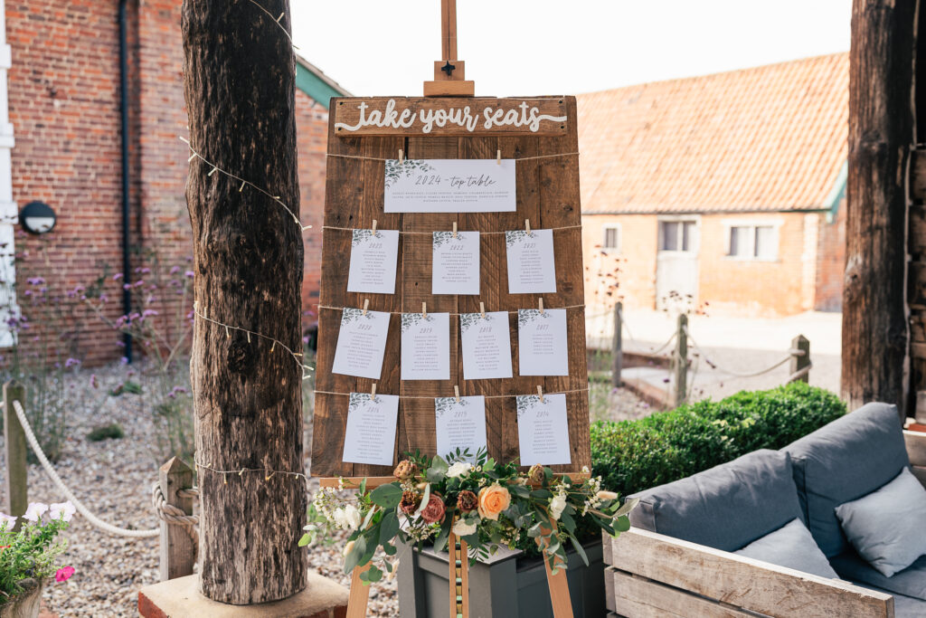 A rustic seating chart displayed on a wooden easel with "Take your seats" written at the top. It features neatly arranged names on paper cards, adorned with floral decorations and surrounded by flowers at the base. The setting is outdoors.