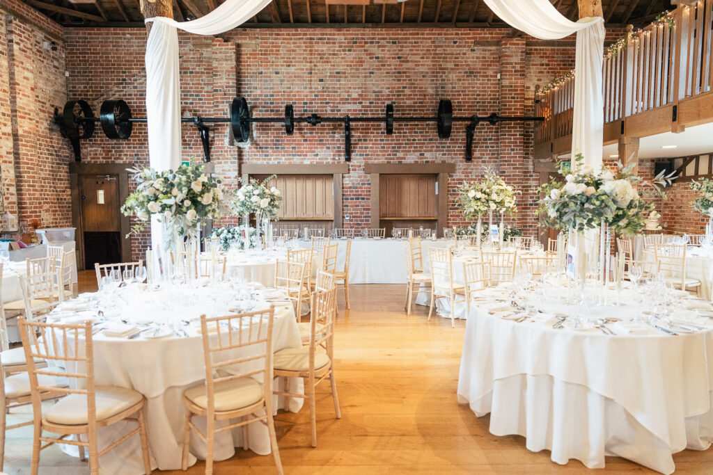 A beautifully decorated indoor wedding reception with round tables set with white tablecloths, elegant place settings, and tall floral centerpieces in a rustic venue with exposed brick walls and wooden beams.