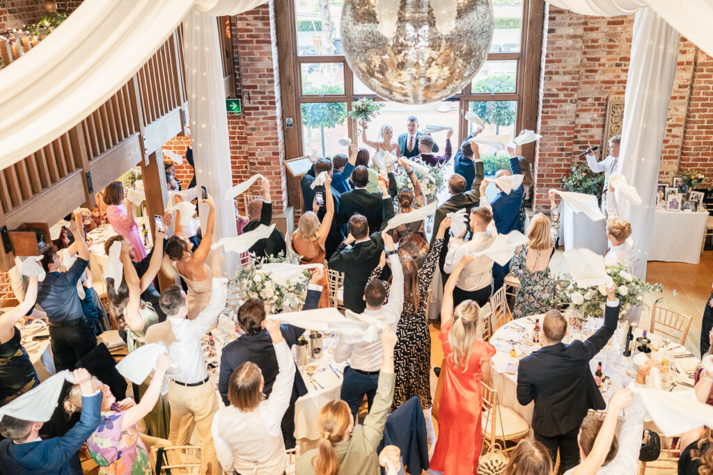 A group of elegantly dressed people is celebrating at an indoor event. They are all standing around decorated tables, raising white napkins in the air. A large disco ball hangs from the ceiling, and a couple stands at the front near large windows.