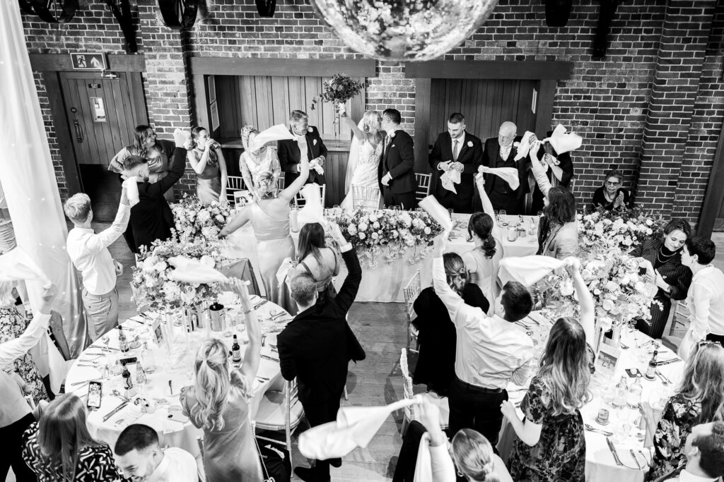 A black and white photo of a wedding reception. Guests seated at tables wave napkins as a couple kisses at the head table adorned with flowers. An exposed brick wall and a large mirror ball are visible in the background.