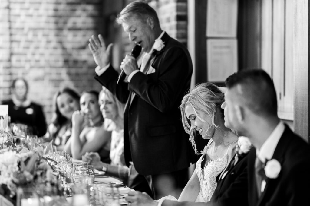 A black and white photo of a wedding reception. A man is giving a speech at a table, holding a microphone. The bride, smiling, is seated beside the groom, who is looking at her. Guests are seated alongside, attentively listening.