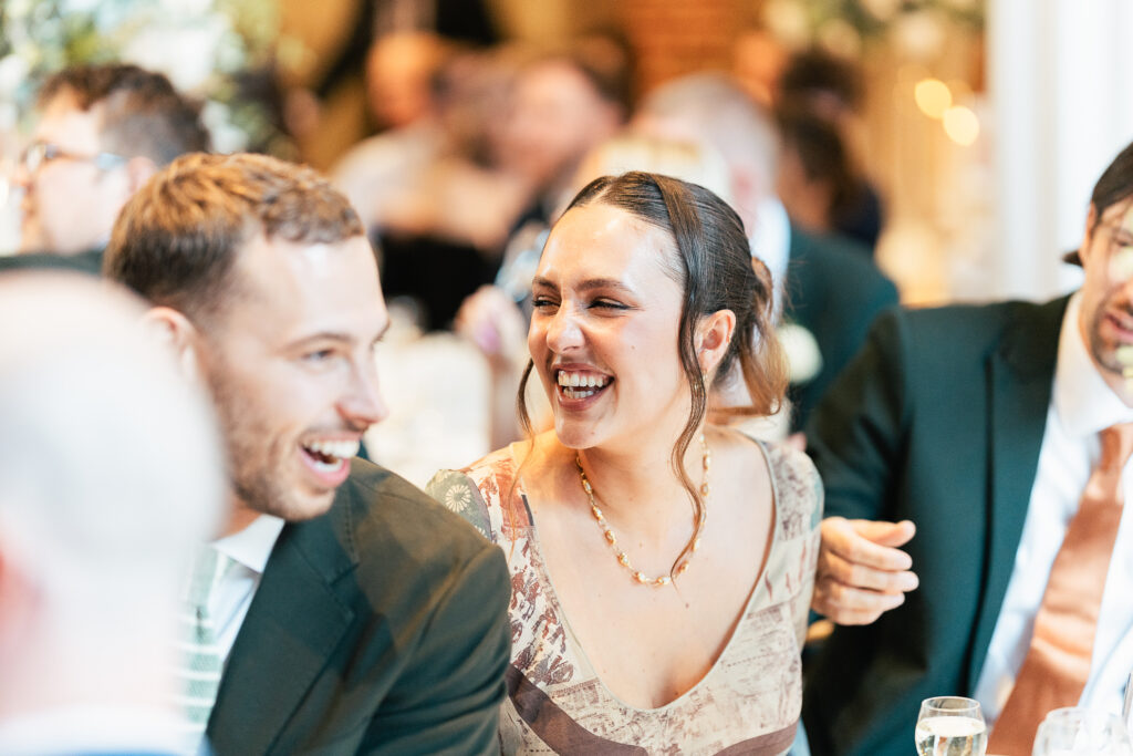 A woman and man are sitting together at an event, laughing and enjoying themselves. The background is filled with other people seated, and the atmosphere appears festive and lively.