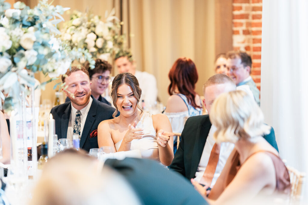 A woman in a white dress is laughing and gesturing while seated at a table during a formal event. She is surrounded by other people in formal attire, with floral decorations in the background. Everyone appears to be enjoying themselves.