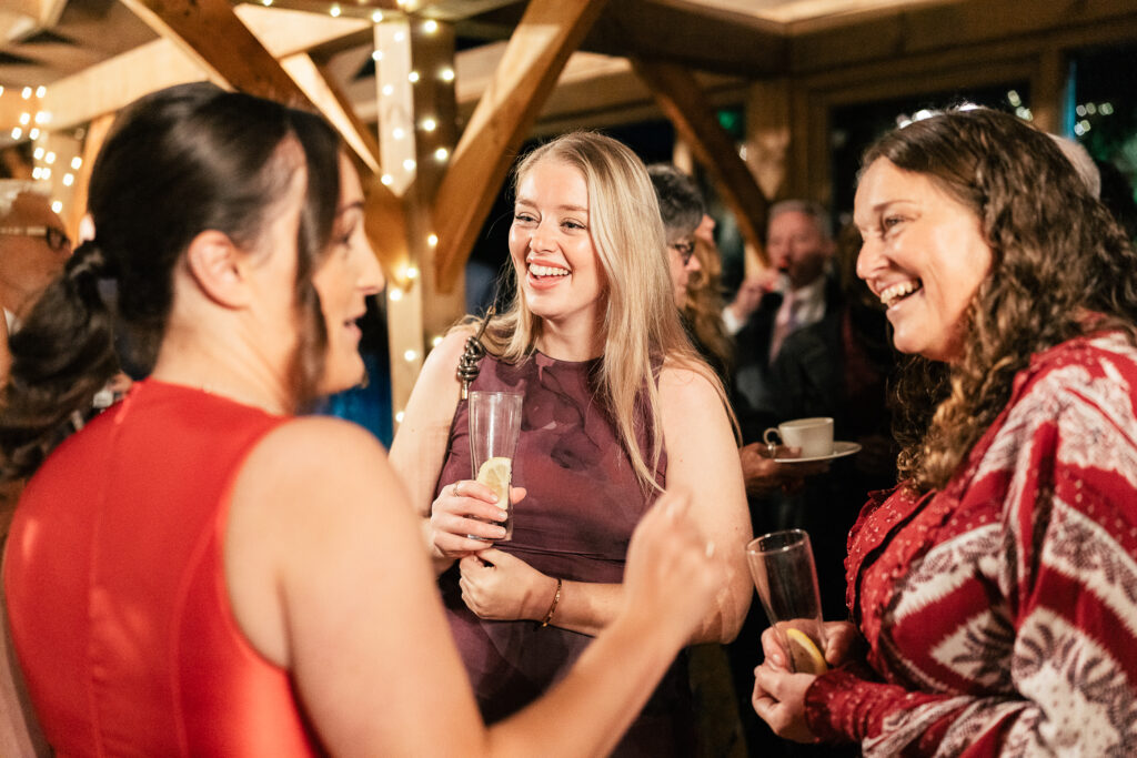 Three women in festive attire are smiling and holding drinks at a lively indoor event. The room features wooden beams and string lights, with several people gathered in the background.