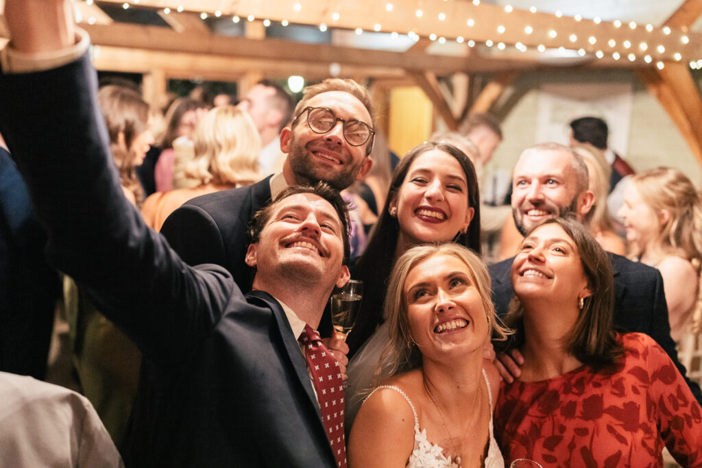 A group of people taking a cheerful selfie at a lively indoor party, surrounded by other guests. The setting features wooden beams and string lights, creating a warm, festive atmosphere.