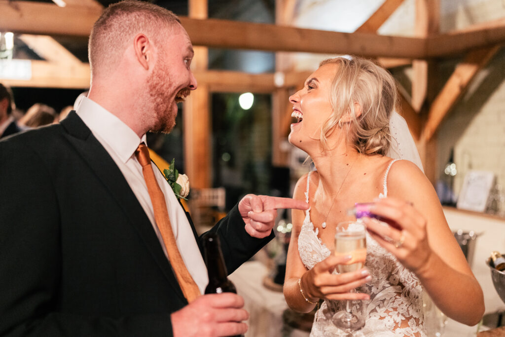 A bride and groom laugh together in a warmly lit setting. The groom, in a suit and tie, gestures playfully with a bottle. The bride, wearing a lace dress and holding a glass of champagne, smiles broadly. Wooden beams and soft lights are in the background.