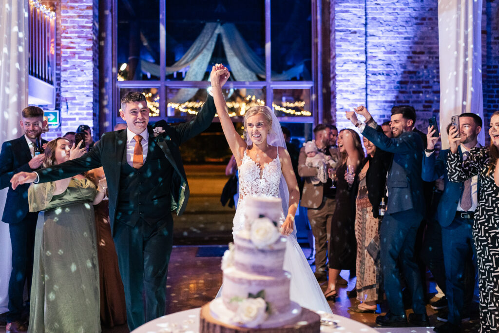 A bride and groom enter a wedding reception hall, holding hands and smiling. Guests stand around, clapping and taking photos. In the foreground is a tiered wedding cake. The setting has blue lighting and brick walls.