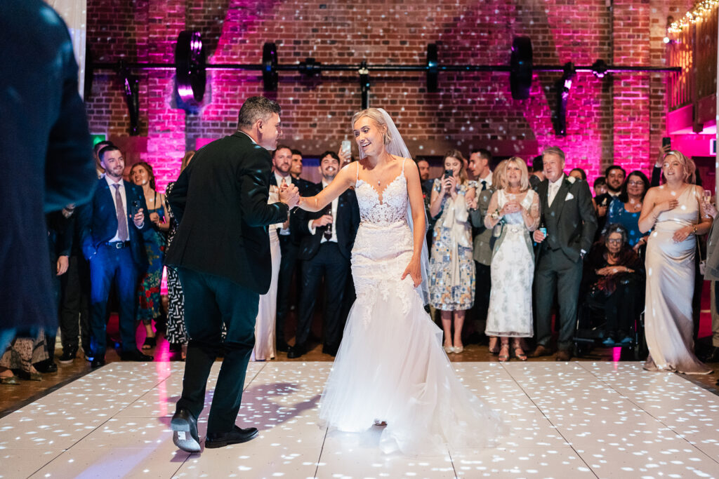 A bride and groom dance on a white floor under colorful lights, surrounded by cheering guests. The brick wall in the background adds to the festive atmosphere, with guests clapping and taking photos of the couple.