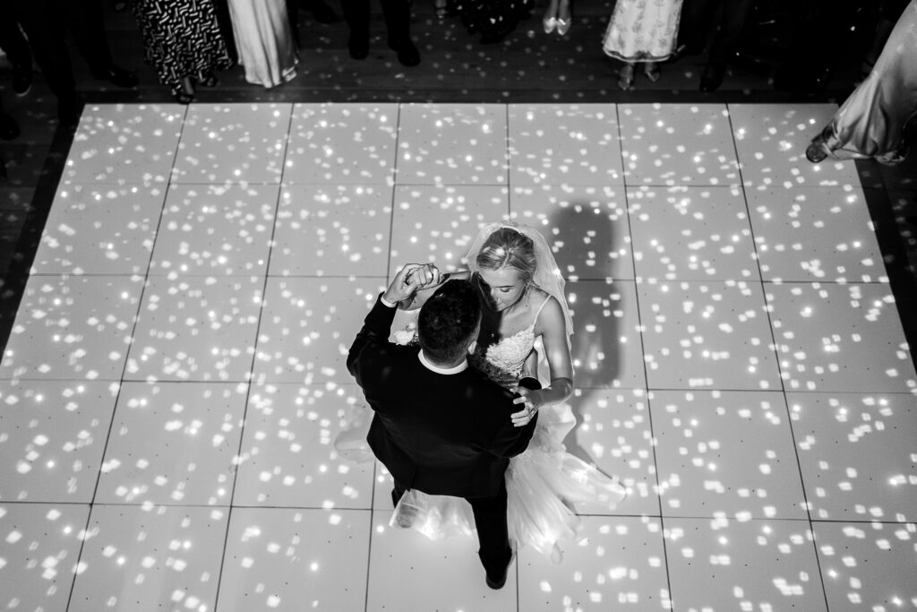 A black-and-white photo of a bride and groom dancing on a checkered dance floor. Sparkling lights create a festive atmosphere. The bride wears a white gown, and the groom is in a dark suit. They are seen from above, surrounded by guests.