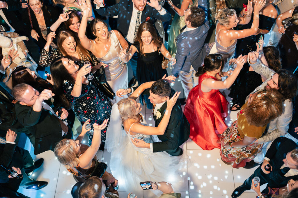 A joyous wedding reception with guests dancing around a bride and groom in the center. The couple shares a dance, surrounded by friends and family celebrating on a lively dance floor. Colorful dresses and suits create a festive atmosphere.